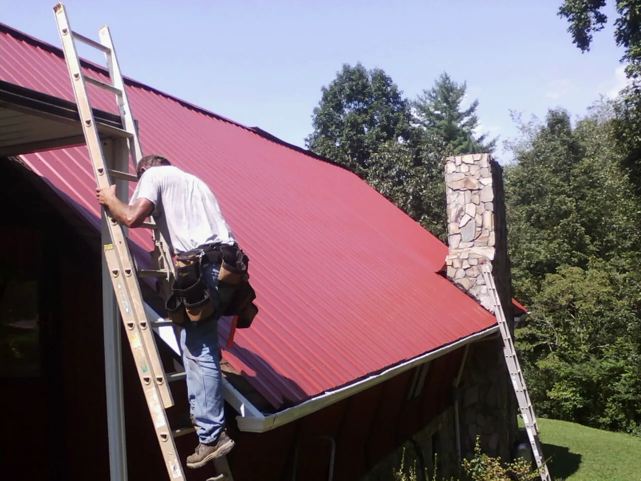 Roofer on a ladder beside a red metal roof and stone chimney — Soffit & Fascia Repair by Roofing Brothers in Paonia